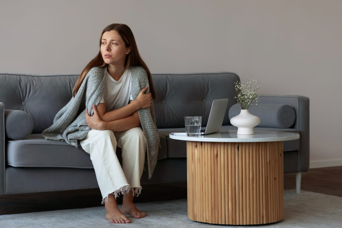 Young woman sitting on a couch wrapped in a blanket, looking worried while holding her arms close to her body in a living room