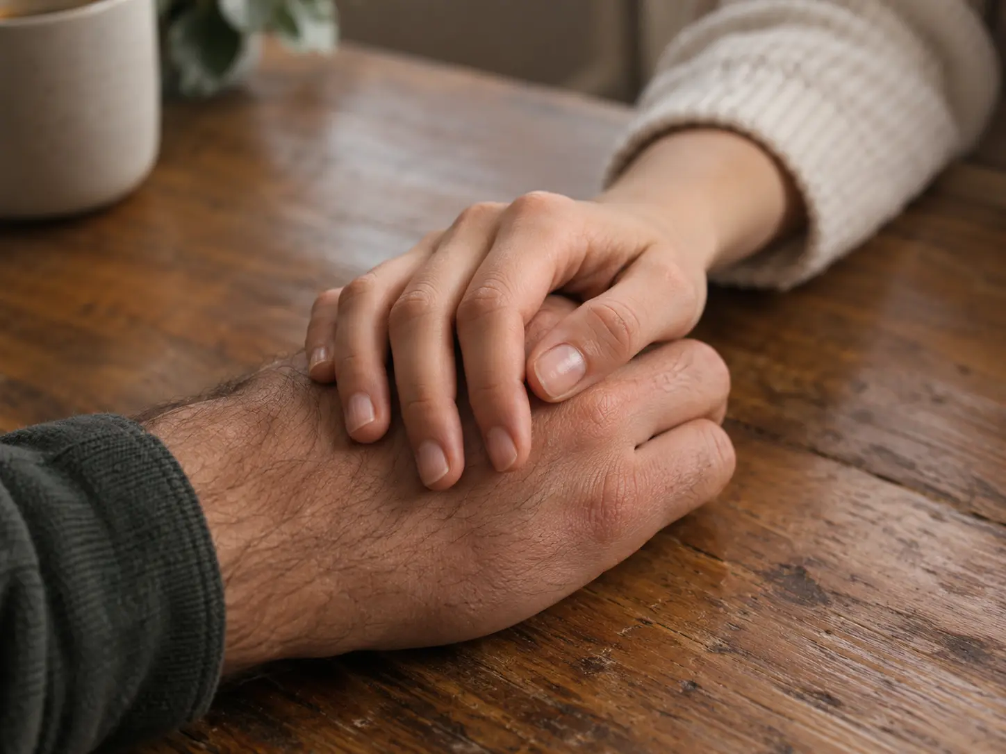 Two hands rest together on a wooden table in a gesture of quiet reassurance.