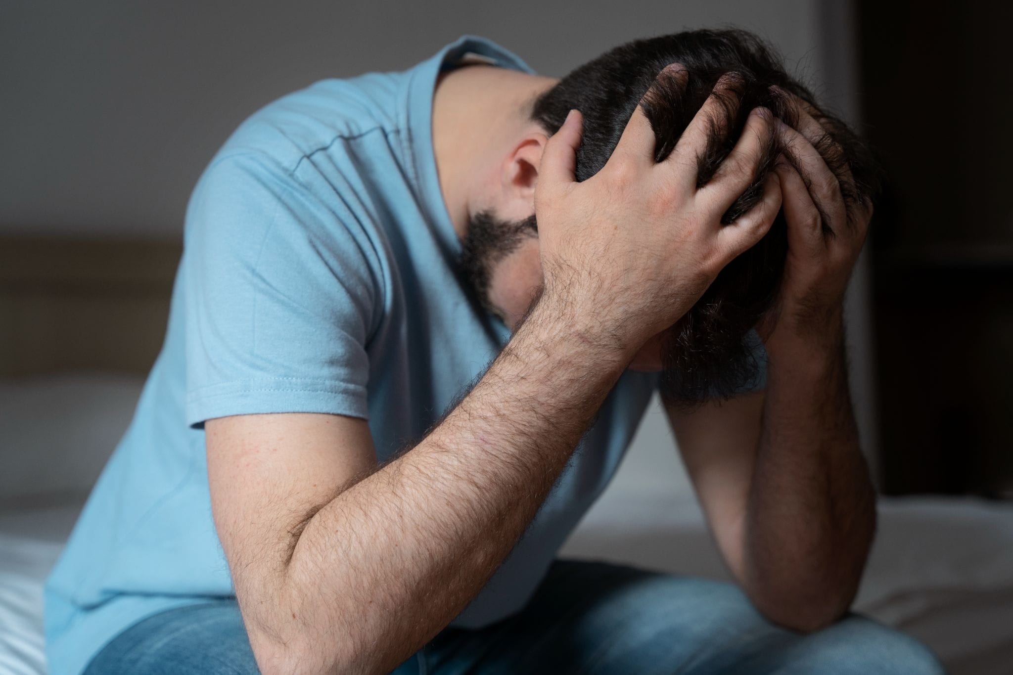 A man sits on the bed, hands on his head, appearing depressed, which is a sign his weed use is now requiring professional help