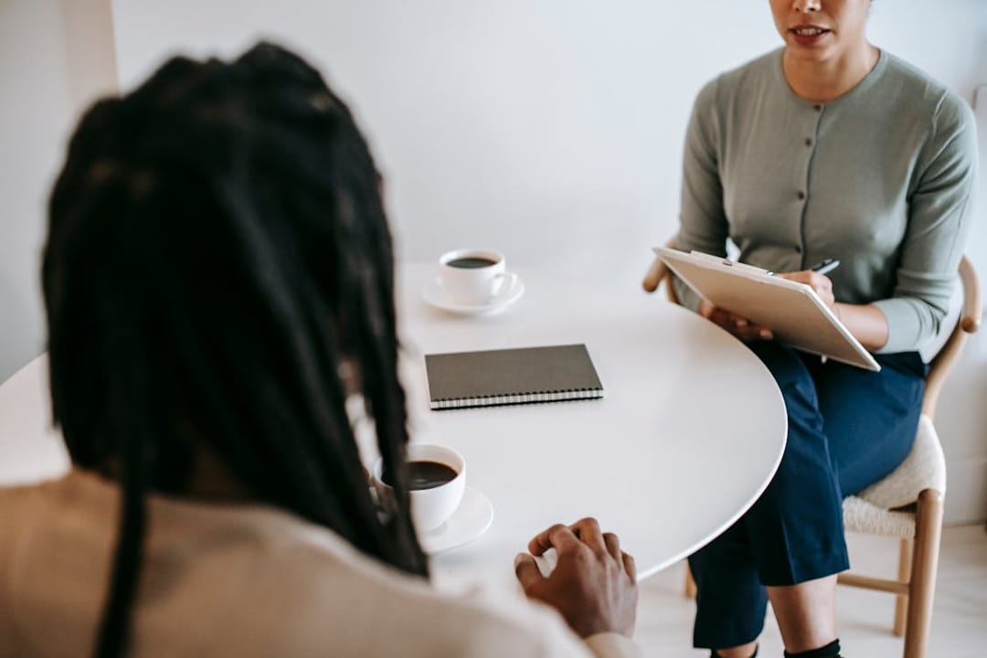 Free Professional ethnic female psychotherapist asking questions to black male patient and taking notes in clipboard while sitting together at table in modern psychotherapy center Stock Photo
